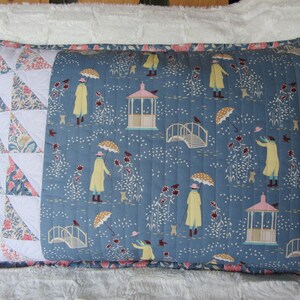 May include: A blue and white pillow with a patchwork design on one side and a repeating pattern of people walking in the rain on the other side. The pattern includes umbrellas, rain, and a gazebo.