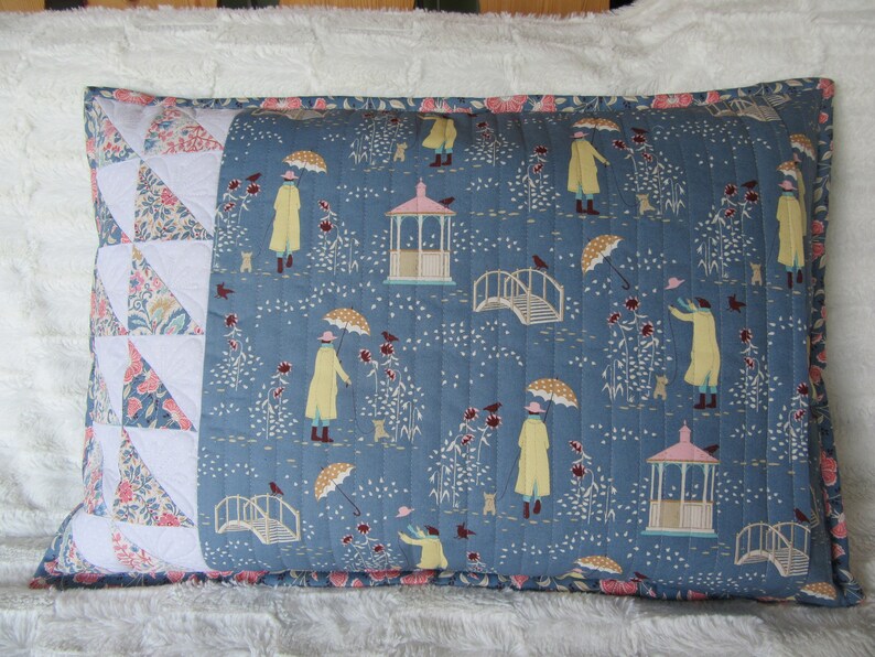 May include: A blue and white pillow with a patchwork design on one side and a repeating pattern of people walking in the rain on the other side. The pattern includes umbrellas, rain, and a gazebo.