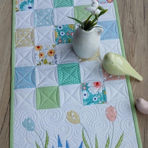 Può includere: Runner da tavolo trapuntato con un design patchwork di quadrati in colori pastello e motivi floreali. Il bordo inferiore presenta tulipani ricamati e altri fiori. Il runner è bordato con un tessuto verde a pois.