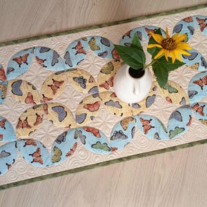 Puede incluir: Un camino de mesa acolchado rectangular con un patrón repetitivo de círculos entrelazados en tela azul claro y amarilla con estampado de mariposas. El fondo es crema con un diseño de costura en espiral y un borde verde fino.