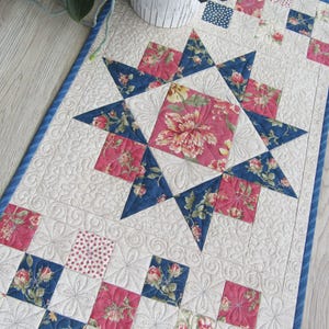 Könnte beinhalten: Gesteppter Tischläufer mit einem zentralen Sterndesign. Der Stern besteht aus blauen Dreiecken und einem zentralen Quadrat mit floralem Muster. Quadrate mit floralen und gepunkteten Mustern in Rosa und Blau umgeben den Stern. Der Läufer hat einen blauen Rand.