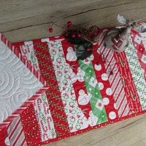 May include: A festive Christmas table runner with a patchwork design. The runner features red and white patterns with candy canes, snowmen, and polka dots. The runner is accented with a red border and white quilted sections. A decorative reindeer figurine and berries are on top.