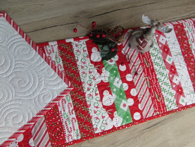 May include: A festive Christmas table runner with a patchwork design. The runner features red and white patterns with candy canes, snowmen, and polka dots. The runner is accented with a red border and white quilted sections. A decorative reindeer figurine and berries are on top.