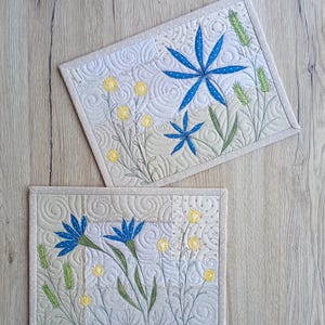 Könnte beinhalten: Zwei rechteckige, gesteppte Tischsets mit floralen Designs. Die Tischsets zeigen blau, gelb und grün gestickte Blumen und Blätter auf cremefarbenem Hintergrund. Die Ränder sind mit einem gepunkteten Rand versehen.