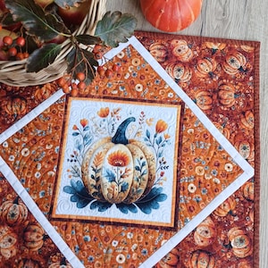 May include: A square quilted table topper featuring a central white panel with a pumpkin and floral illustration, surrounded by a diamond-shaped white border and an outer fabric print of orange pumpkins and small flowers on a dark background.