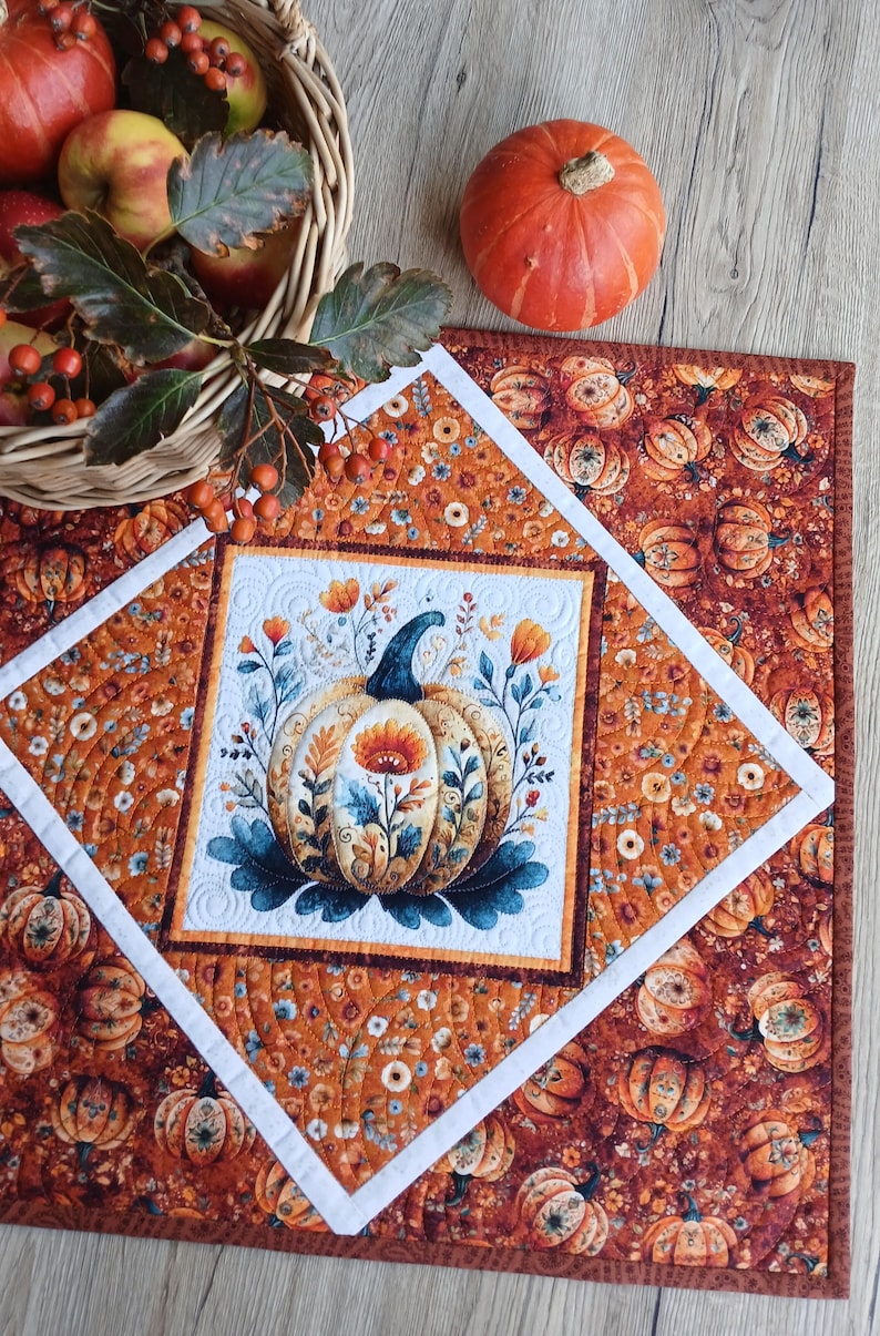 May include: A square quilted table topper featuring a central white panel with a pumpkin and floral illustration, surrounded by a diamond-shaped white border and an outer fabric print of orange pumpkins and small flowers on a dark background.