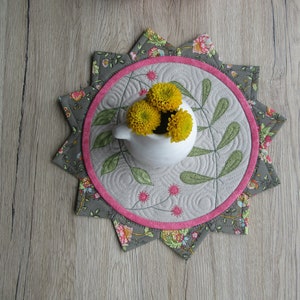 K&ouml;nnte beinhalten: Ein grauer und rosa gesteppter Tischl&auml;ufer mit einem Sternendesign. Die Mitte des L&auml;ufers zeigt eine wei&szlig;e Keramikvase mit gelben Blumen.