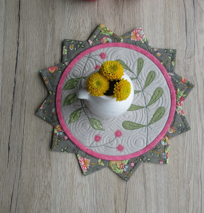 K&ouml;nnte beinhalten: Ein grauer und rosa gesteppter Tischl&auml;ufer mit einem Sternendesign. Die Mitte des L&auml;ufers zeigt eine wei&szlig;e Keramikvase mit gelben Blumen.