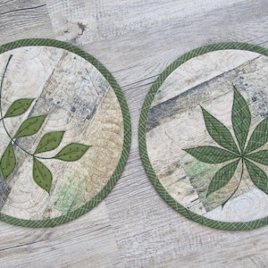 Könnte beinhalten: Zwei runde Stoffuntersetzer mit grünen Blattmotiven. Einer zeigt einen Zweig mit Blättern, der andere ein mehrlappiges Blatt. Die Untersetzer haben einen grünen Rand und einen Hintergrund in Holzoptik.