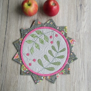 Può includere: Un sottopentola in tessuto grigio e floreale con un bordo cucito rosa e un design trapuntato di foglie verdi e fiori rosa.