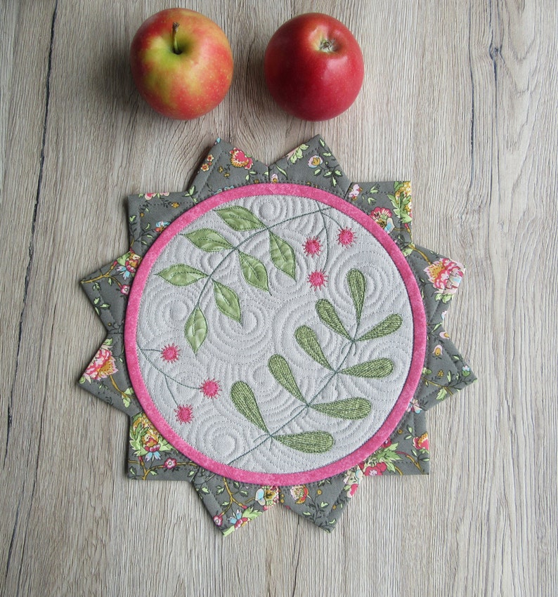K&ouml;nnte beinhalten: Ein grauer und floral gemusterter Stoffuntersetzer mit einem rosa gestickten Rand und einem gesteppten Design aus gr&uuml;nen Bl&auml;ttern und rosa Bl&uuml;ten.
