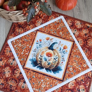 May include: A square quilted table mat featuring a central pumpkin illustration surrounded by floral motifs, framed by a white border and an outer layer of orange fabric with a repeating pumpkin and flower pattern, with a basket of apples nearby.