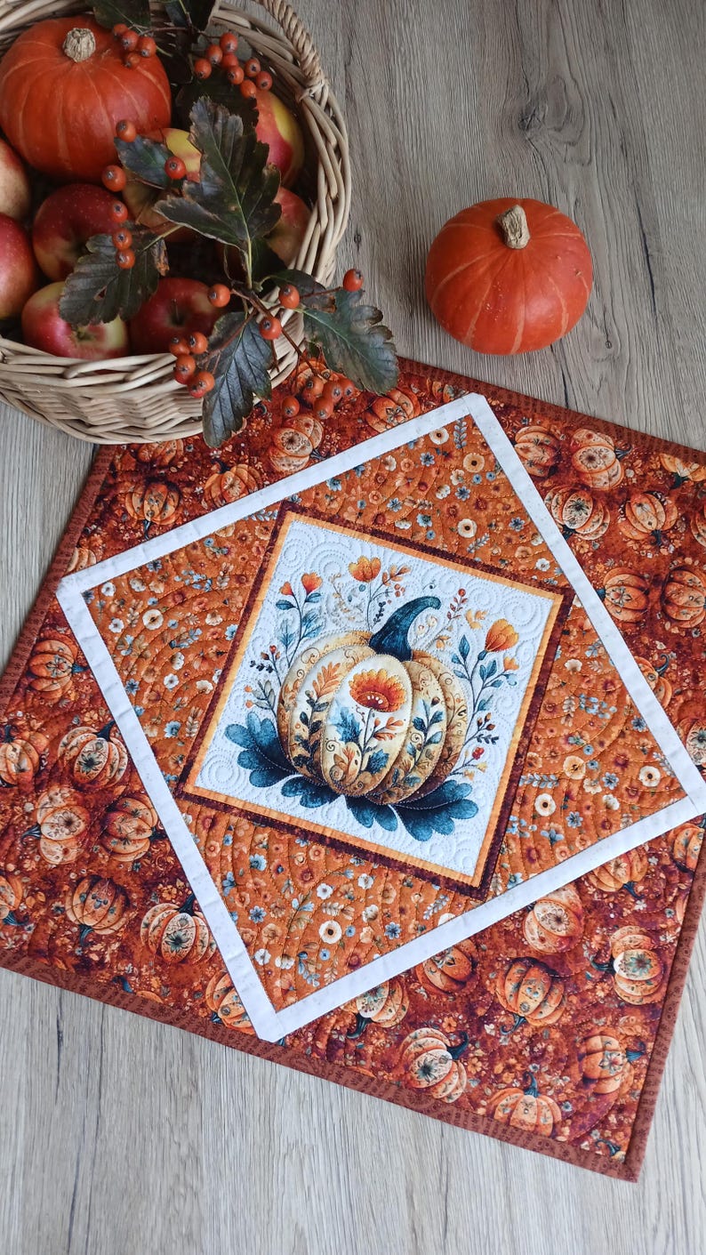 May include: A square quilted table mat featuring a central pumpkin illustration surrounded by floral motifs, framed by a white border and an outer layer of orange fabric with a repeating pumpkin and flower pattern, with a basket of apples nearby.