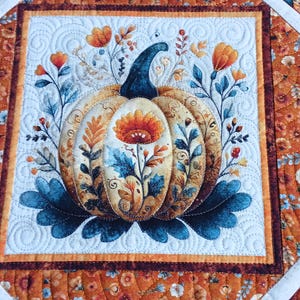 May include: A quilted fabric runner featuring a central square with a pumpkin illustration surrounded by floral motifs in shades of orange, blue, and cream. The border displays a repeating pumpkin and flower pattern with white diagonal fabric strips.