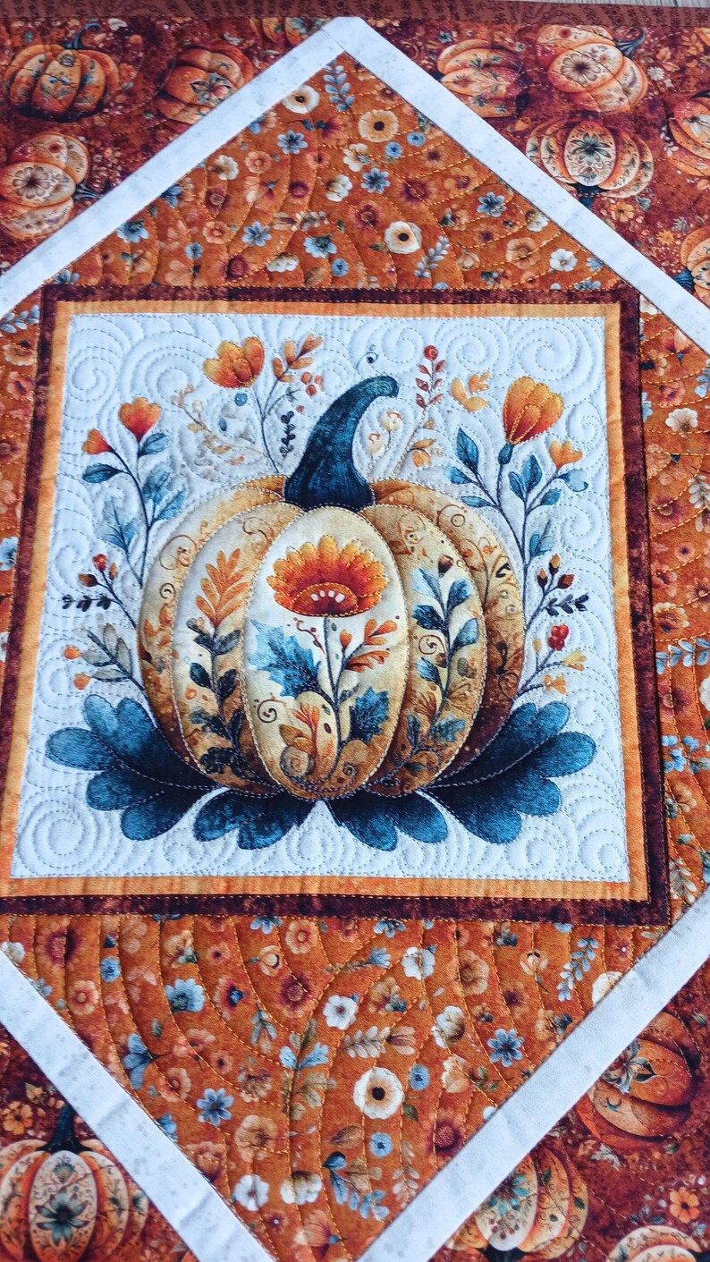 May include: A quilted fabric runner featuring a central square with a pumpkin illustration surrounded by floral motifs in shades of orange, blue, and cream. The border displays a repeating pumpkin and flower pattern with white diagonal fabric strips.
