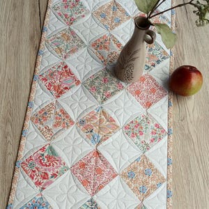 Könnte beinhalten: Ein rechteckiger, gesteppter Tischläufer mit einem sich wiederholenden Muster aus rautenförmigen, floralen Stoffstücken in Rosa-, Blau- und Orangetönen, durchsetzt mit weißen, gesteppten Quadraten, die mit gestickten Blütenblattmotiven verziert sind.