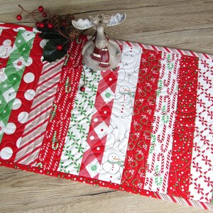 May include: A Christmas-themed table runner with a patchwork design. The runner features alternating stripes of red, white, and green fabrics with patterns including snowmen, candy canes, and polka dots. A decorative moose figurine sits on top.