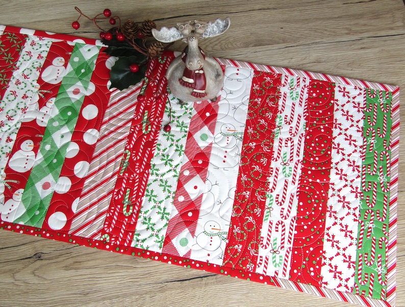 May include: A Christmas-themed table runner with a patchwork design. The runner features alternating stripes of red, white, and green fabrics with patterns including snowmen, candy canes, and polka dots. A decorative moose figurine sits on top.