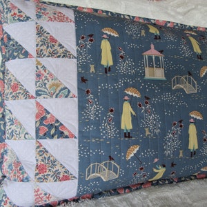 May include: A blue and white pillow with a floral pattern and a white border. The pillow features a repeating pattern of people walking under umbrellas, a bridge, and a gazebo.
