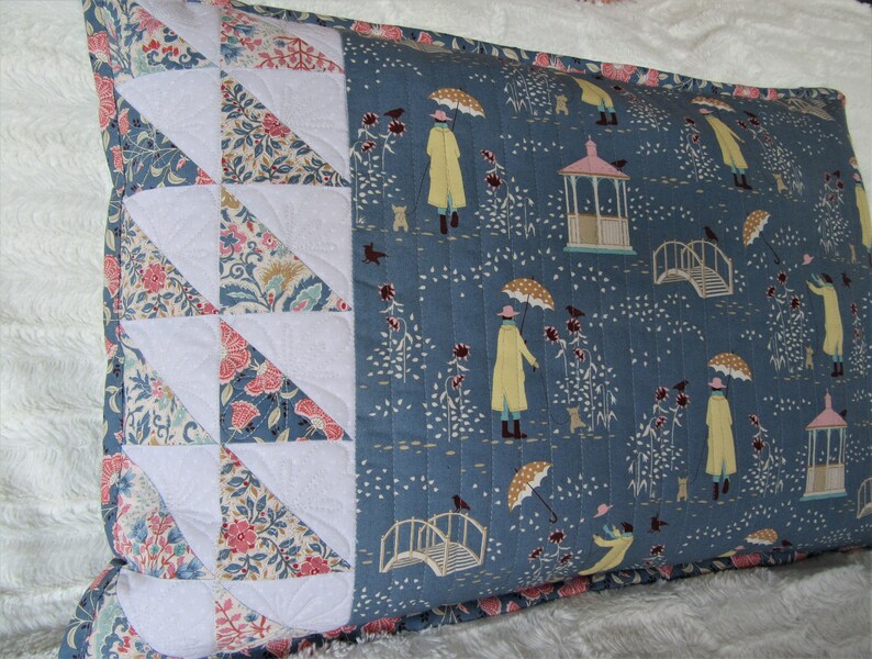 May include: A blue and white pillow with a floral pattern and a white border. The pillow features a repeating pattern of people walking under umbrellas, a bridge, and a gazebo.