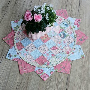 Op de afbeelding: Een bloemen gewatteerde placemat in roze en blauw, met een centraal cirkelvormig ontwerp en een stervorm. Een kleine roze pot met roze rozen en witte bloemen staat erop. De placemat is gemaakt van stof met een patchwork ontwerp.