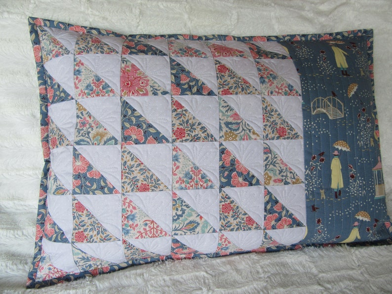 May include: A decorative pillow with a patchwork design featuring a variety of floral patterns in shades of pink, blue, and white. The pillow is quilted with a white fabric and has a blue fabric border. The back of the pillow features a blue fabric with a repeating pattern of people walking in the rain.