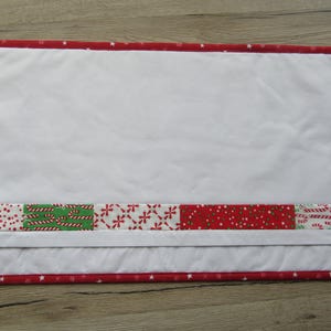 May include: Rectangular white quilted placemat with a red border featuring white stars. A band of Christmas-themed fabric with candy canes, dots, and snowflakes accents the bottom. The placemat is designed for festive table settings.