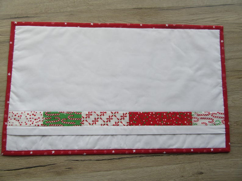 May include: Rectangular white quilted placemat with a red border featuring white stars. A band of Christmas-themed fabric with candy canes, dots, and snowflakes accents the bottom. The placemat is designed for festive table settings.