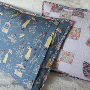 May include: Two decorative pillow covers. One is blue with a white and yellow pattern of people walking in a garden with umbrellas. The other is white with a patchwork design of floral fabric squares.