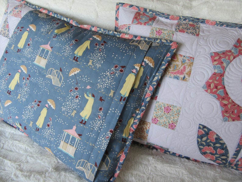 May include: Two decorative pillow covers. One is blue with a white and yellow pattern of people walking in a garden with umbrellas. The other is white with a patchwork design of floral fabric squares.
