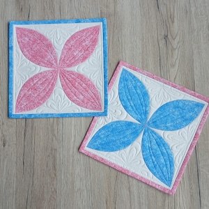 Könnte beinhalten: Zwei quadratische, gesteppte Untersetzer mit floralem Design. Einer hat rosa Blütenblätter mit blauem Rand, der andere blaue Blütenblätter mit rosa Rand. Beide haben weiße Zentren mit dekorativen Nähten.