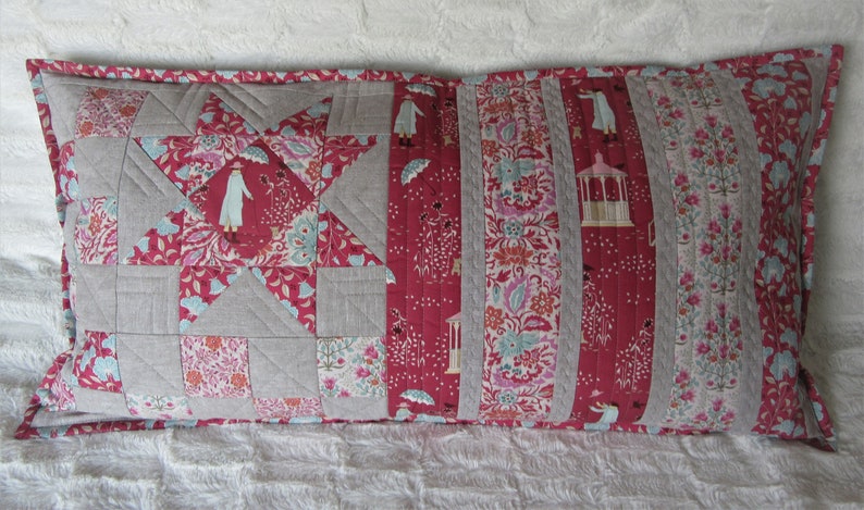 May include: A long rectangular pillow with a patchwork design. The pillow is made of various fabrics in shades of red, gray, and white. The front of the pillow features a star design with a floral pattern. The back of the pillow is a solid red fabric.