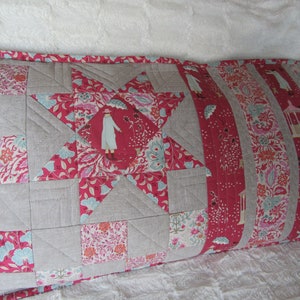 May include: A long rectangular pillow with a patchwork design. The pillow is made of a variety of fabrics, including a light gray linen and a red floral print. The pillow is quilted with a star pattern in the center.