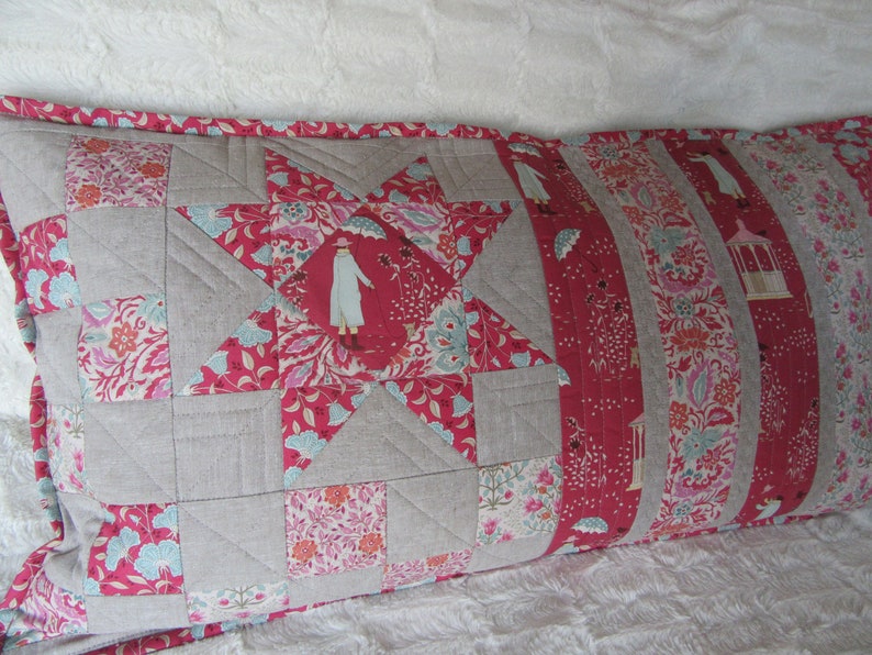 May include: A long rectangular pillow with a patchwork design. The pillow is made of a variety of fabrics, including a light gray linen and a red floral print. The pillow is quilted with a star pattern in the center.
