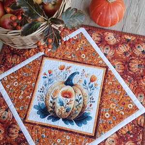 May include: A square quilted fabric mat featuring a central white panel with a printed pumpkin and floral design, surrounded by an orange border with a repeating pumpkin and flower pattern, accented by a white frame and intricate stitching.