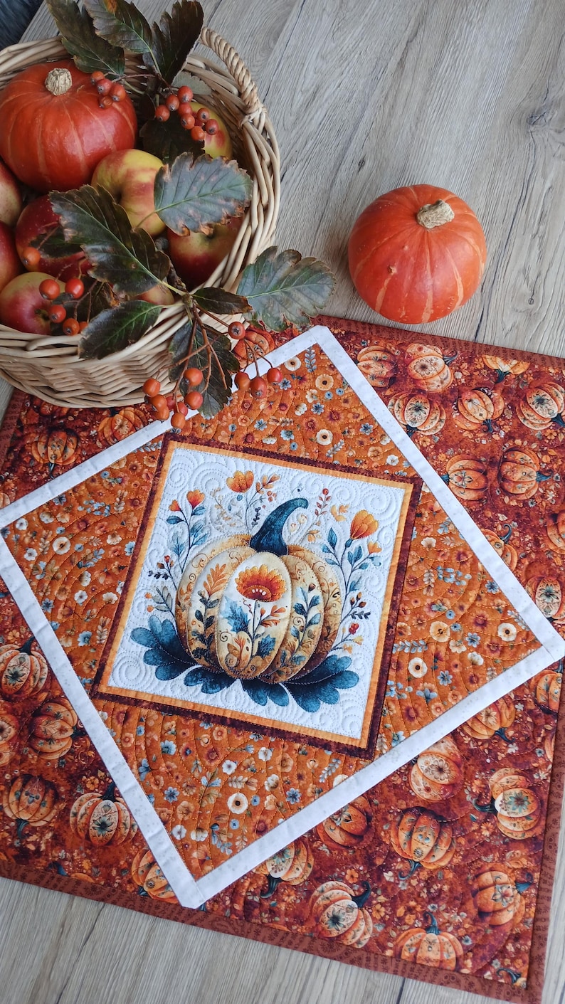 May include: A square quilted fabric mat featuring a central white panel with a printed pumpkin and floral design, surrounded by an orange border with a repeating pumpkin and flower pattern, accented by a white frame and intricate stitching.
