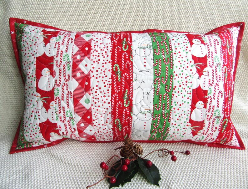 May include: Rectangular Christmas pillow with a red border. The pillow features vertical stripes with patterns of snowmen, candy canes, and red and white polka dots. The pillow is decorated with a pine cone, holly leaves, and red berries.