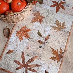 May include: A rectangular quilted table runner featuring a cream-colored background with vintage script text and various brown, orange, and green fabric maple leaf appliques. The runner has intricate swirl stitching and a brown patterned border.