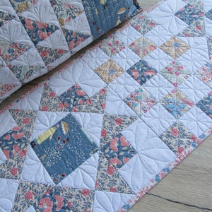 May include: A white quilted table runner with a floral pattern in shades of blue, pink, and brown. The runner is made of fabric squares sewn together in a diamond pattern.