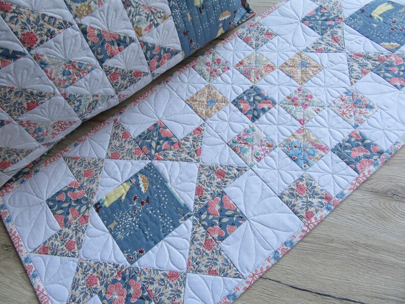 May include: A white quilted table runner with a floral pattern in shades of blue, pink, and brown. The runner is made of fabric squares sewn together in a diamond pattern.