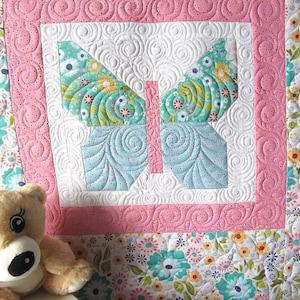 Könnte beinhalten: Eine rosa-weiße Babydecke mit einem blauen und grünen Schmetterlingsmuster. Die Decke ist mit einem Wirbelmuster gesteppt und hat einen floralen Rand.