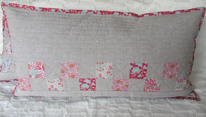 May include: A long rectangular pillow with a light gray linen fabric cover. The pillow has a pink and white floral patterned border and a bottom row of patchwork squares in pink, white, and blue floral patterns.
