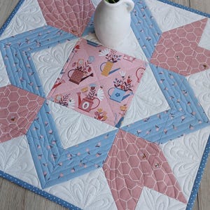 Puede incluir: Camino de mesa acolchado con un diseño floral y geométrico. El camino de mesa presenta un cuadrado central con regaderas y flores, rodeado de formas geométricas azules, blancas y rosas. Un pequeño jarrón blanco con flores encima.