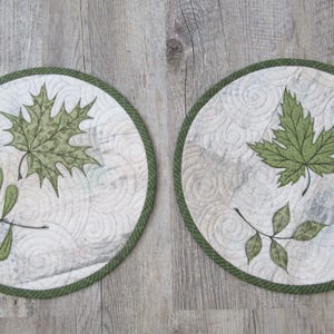 Könnte beinhalten: Zwei runde, gesteppte Tischsets mit weißem Hintergrund und grünen Blattmotiven. Jedes Tischset hat einen grünen Rand. Eines zeigt ein Ahornblatt und das andere kleinere Blätter.