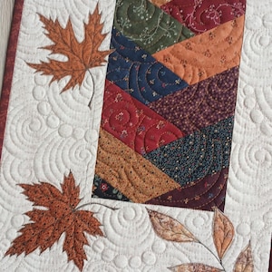 May include: A rectangular quilted table runner featuring a central column of diagonal fabric strips in autumn colors like orange, red, green, and navy blue. The cream-colored border displays intricate swirl quilting and appliqued maple leaves.