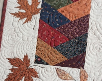 May include: A rectangular quilted table runner featuring a central column of diagonal fabric strips in autumn colors like orange, red, green, and navy blue. The cream-colored border displays intricate swirl quilting and appliqued maple leaves.