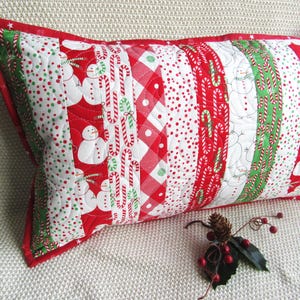 May include: A rectangular Christmas-themed pillow with a red border. The pillow features a patchwork design with alternating stripes of snowmen, candy canes, polka dots, and gingham patterns in red, white, and green.