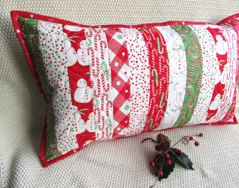 May include: A rectangular Christmas-themed pillow with a red border. The pillow features a patchwork design with alternating stripes of snowmen, candy canes, polka dots, and gingham patterns in red, white, and green.