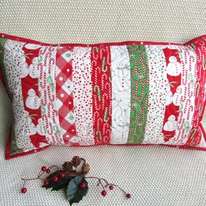 May include: Rectangular Christmas pillow with a red border. The pillow features a patchwork design with alternating stripes of snowmen, candy canes, and red and white patterns. The pillow is on a white surface.