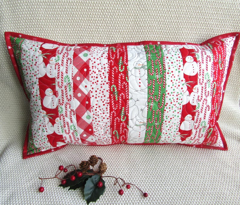 May include: Rectangular Christmas pillow with a red border. The pillow features a patchwork design with alternating stripes of snowmen, candy canes, and red and white patterns. The pillow is on a white surface.